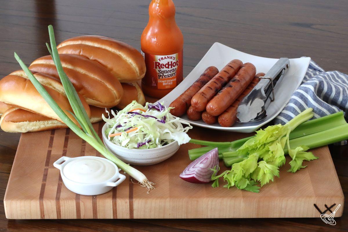 Ingredients on a cutting board including hot dog wieners, hot dog buns, and coleslaw.