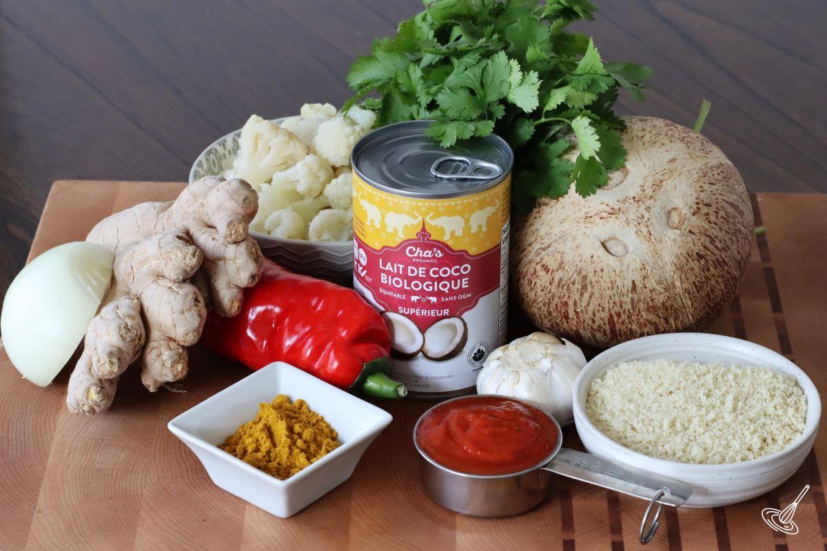 Ingredients on a wooden board, including coconut milk, panko crumbs, cauliflower florets, ginger root, and garlic. 