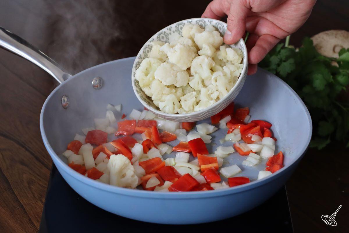 Someone adding cauliflower flaurets to a frying pan with bell pepper and onion. 