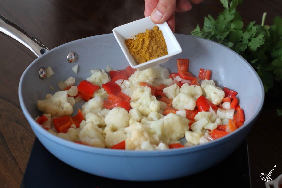 Someone adding curry powder to a large frying pan containing vegetables. 