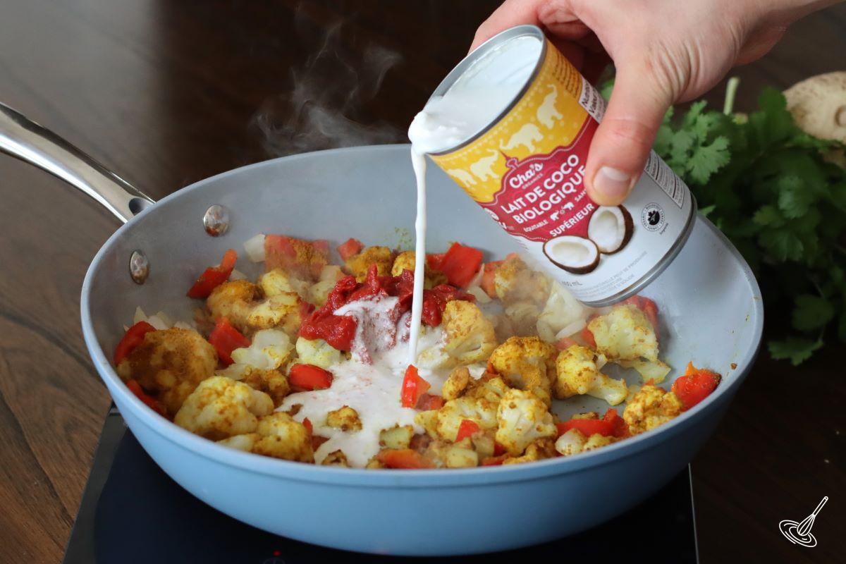 Someone pouring in a can of coconut milk into a frying pan of curry vegetables. 