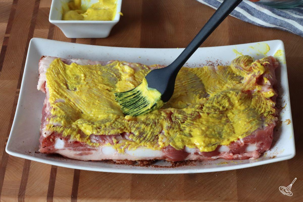 Someone brushing yellow mustard on a rack of ribs. 