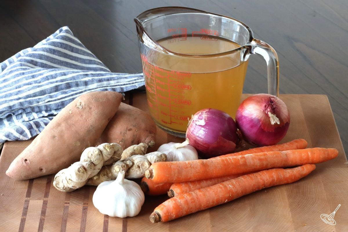 Carrots, sweet potatoes, onion, ginger, and vegetable broth on a cutting board. 