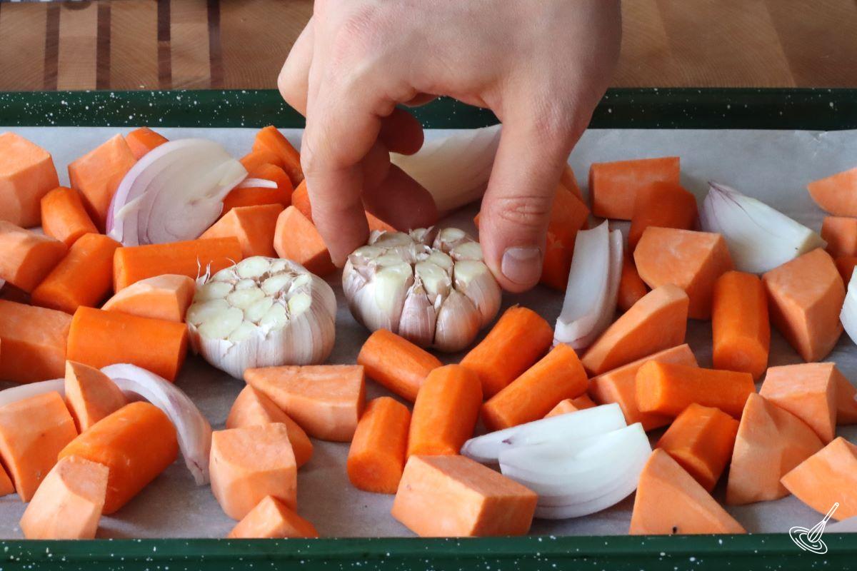 Someone placing a garlic bulb on a baking tray with over vegetables.