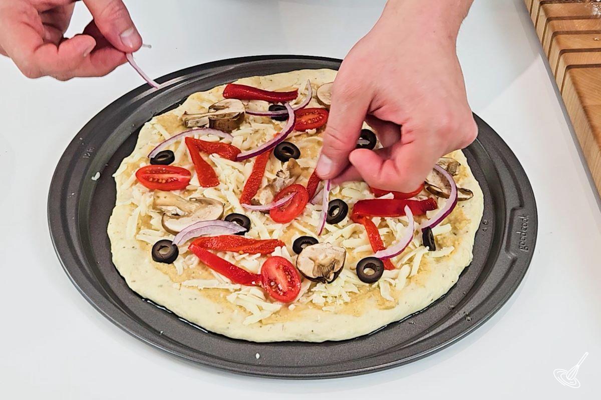 Someone adding vegetables on top of the Hummus Pizza.