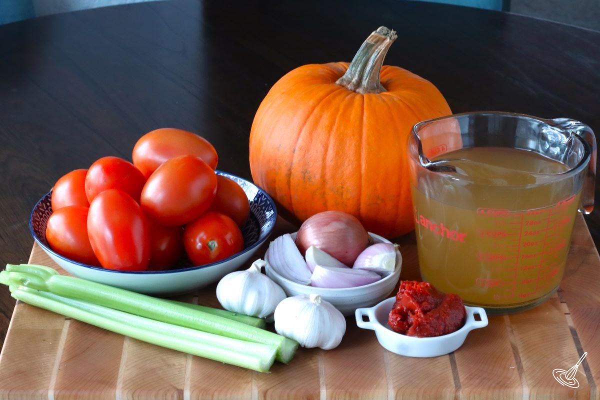 Ingredients on a cutting board including a pumpkin, tomatoes, and onions. 
