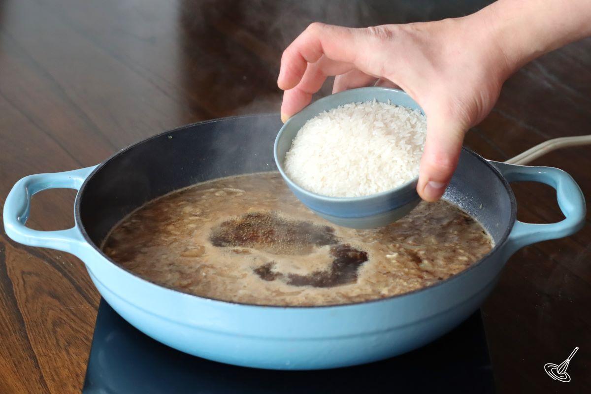 Someone adding rice to a skillet of simmering broth.