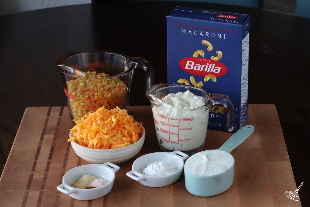 Ingredients on a cutting board, including macaroni, grated cheese, cottage cheese, and Greek yogurt.