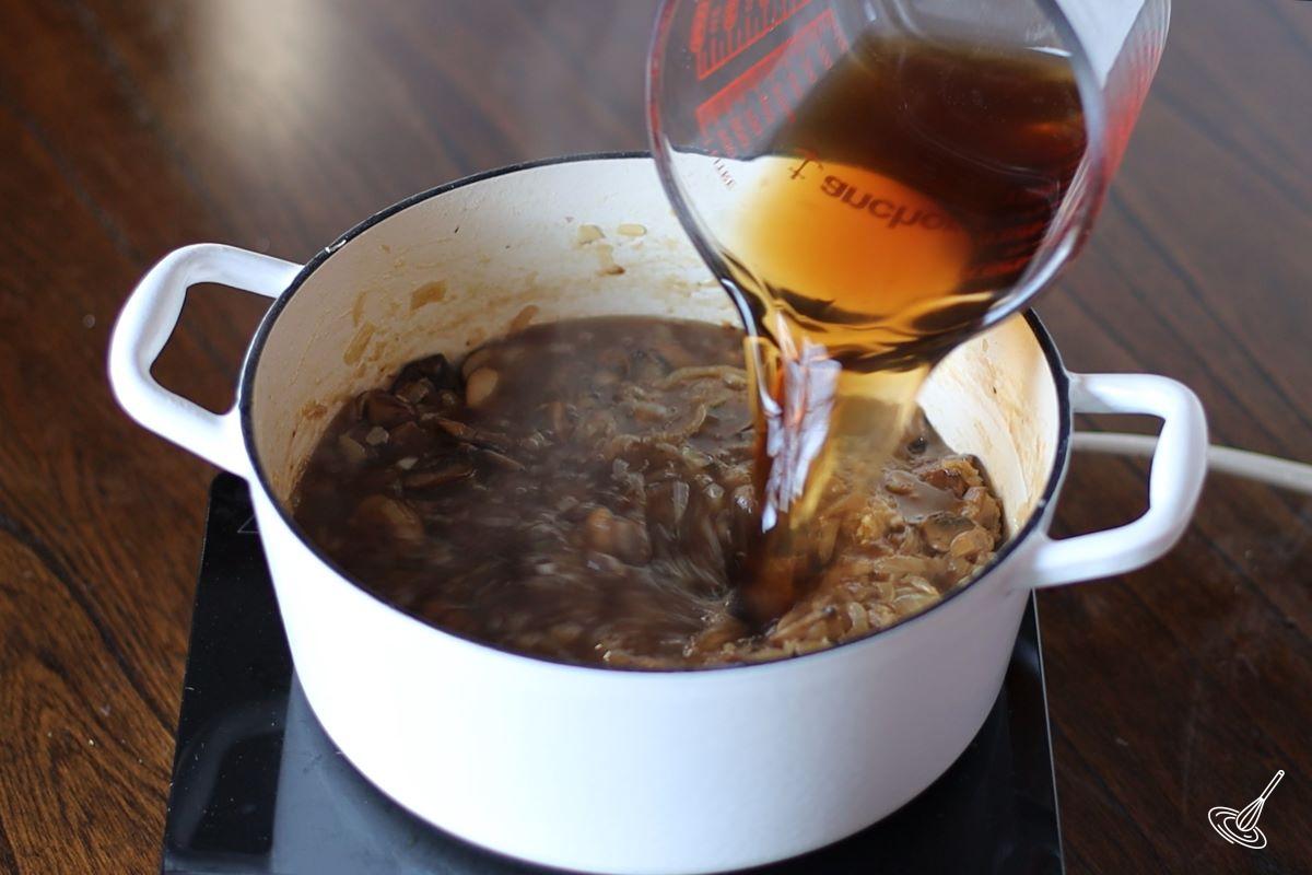 Someone pouring beef broth in a Dutch oven of onion soup.