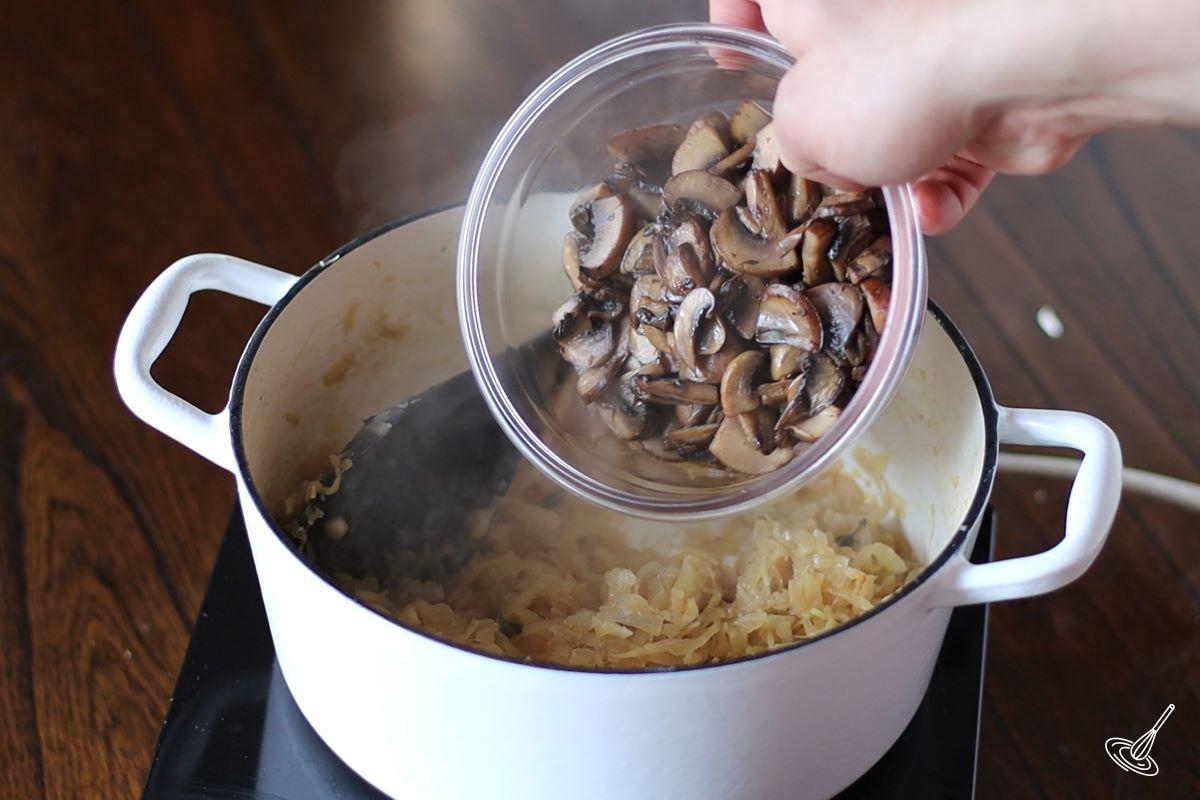 Someone adding cooked mushrooms to a Dutch oven containing caramelized onions.