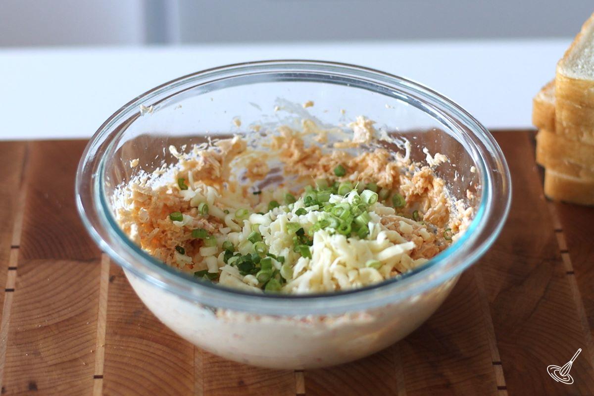 Someone adding sliced green onion and grated cheese to a bowl of crab Rangoon filling. 