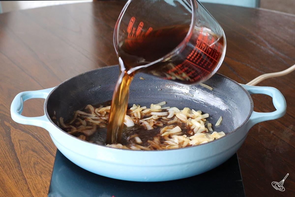 Someone pouring beef broth into a skillet with cooked onions.