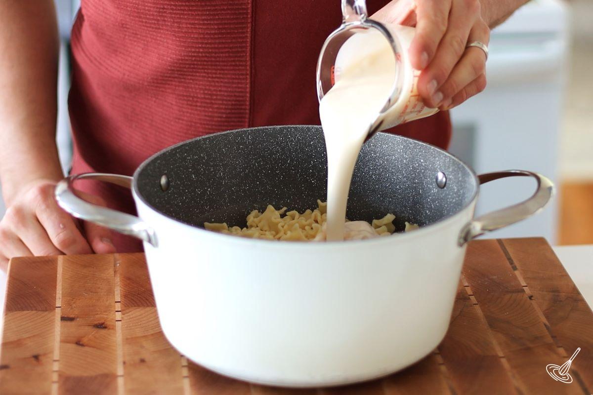 Someone adding alfredo sauce to a pot of cooked pasta.