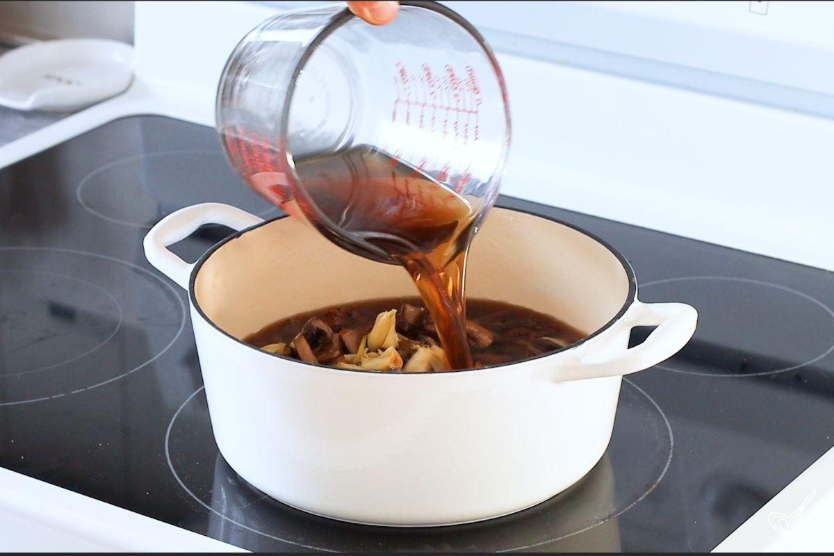 Someone pouring beef broth in a Dutch oven with vegetables.