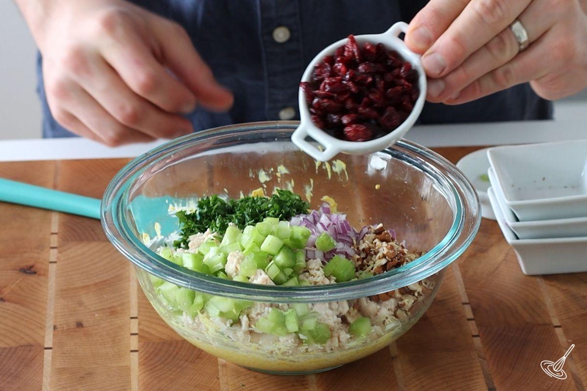 Someone adding craisins to a large bowl of curry chicken salad.