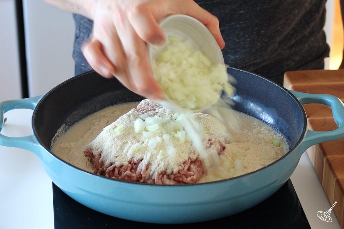 Someone adding chopped onions to a large skillet with ground pork.