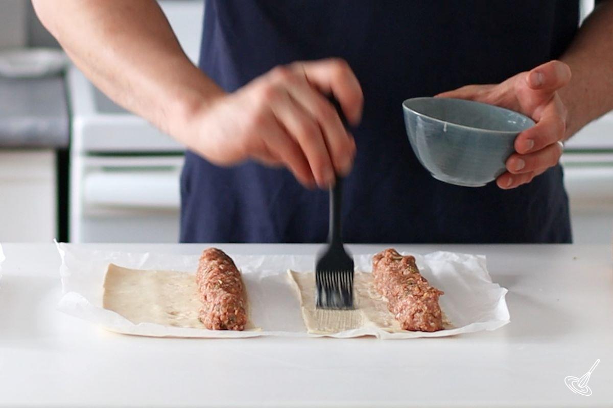 Someone brushing egg wash on the inside of a sausage roll.