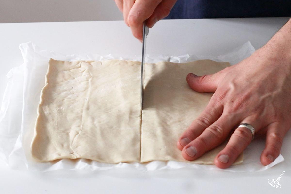 Someone cutting a flat square of puff pastry in half.