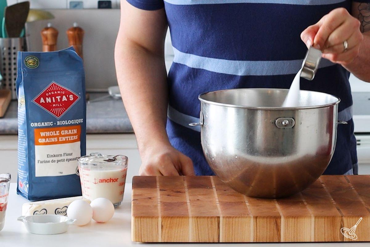 Someone adding sugar to a large mixing bowl.