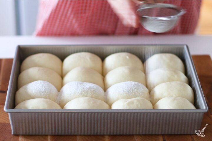 Someone dusting flour over proofed dinner rolls.
