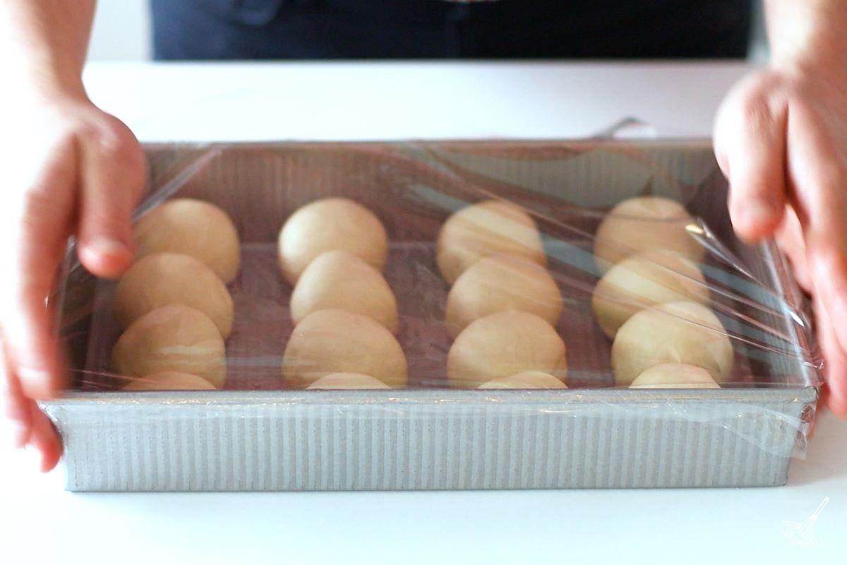 Someone placing plastic wrap over a baking dish of dinner rolls.