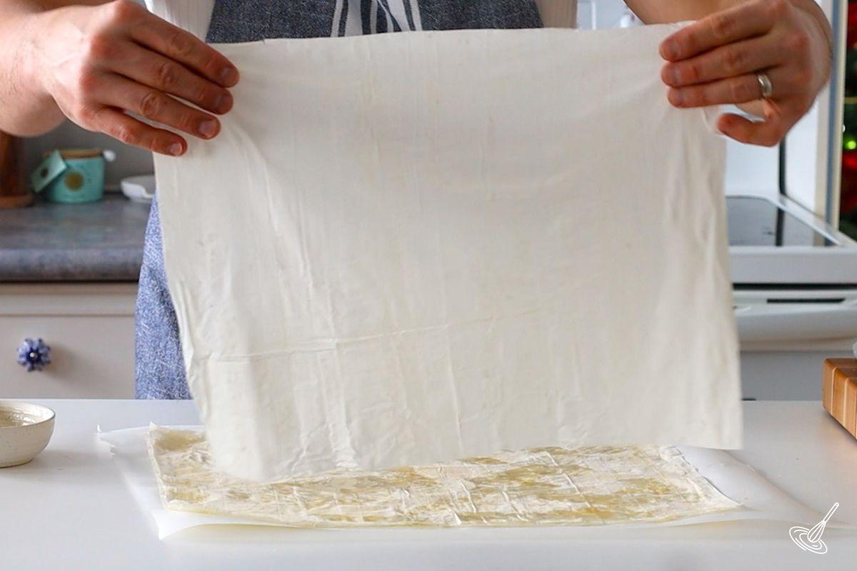 Someone placing a sheet of phyllo dough on top of a a buttered sheet.