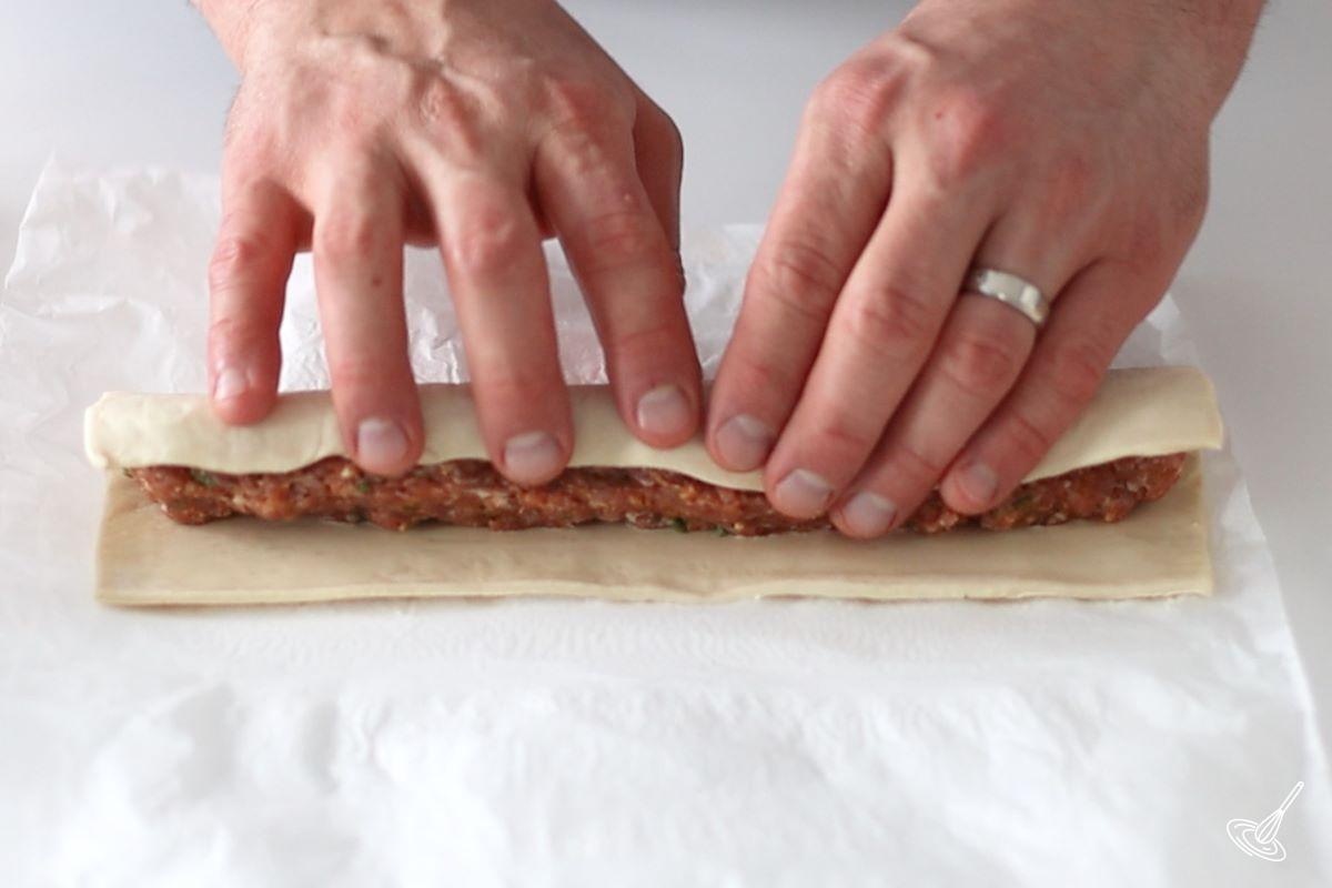 Someone using their hands to encase a log of meat with puff pastry.