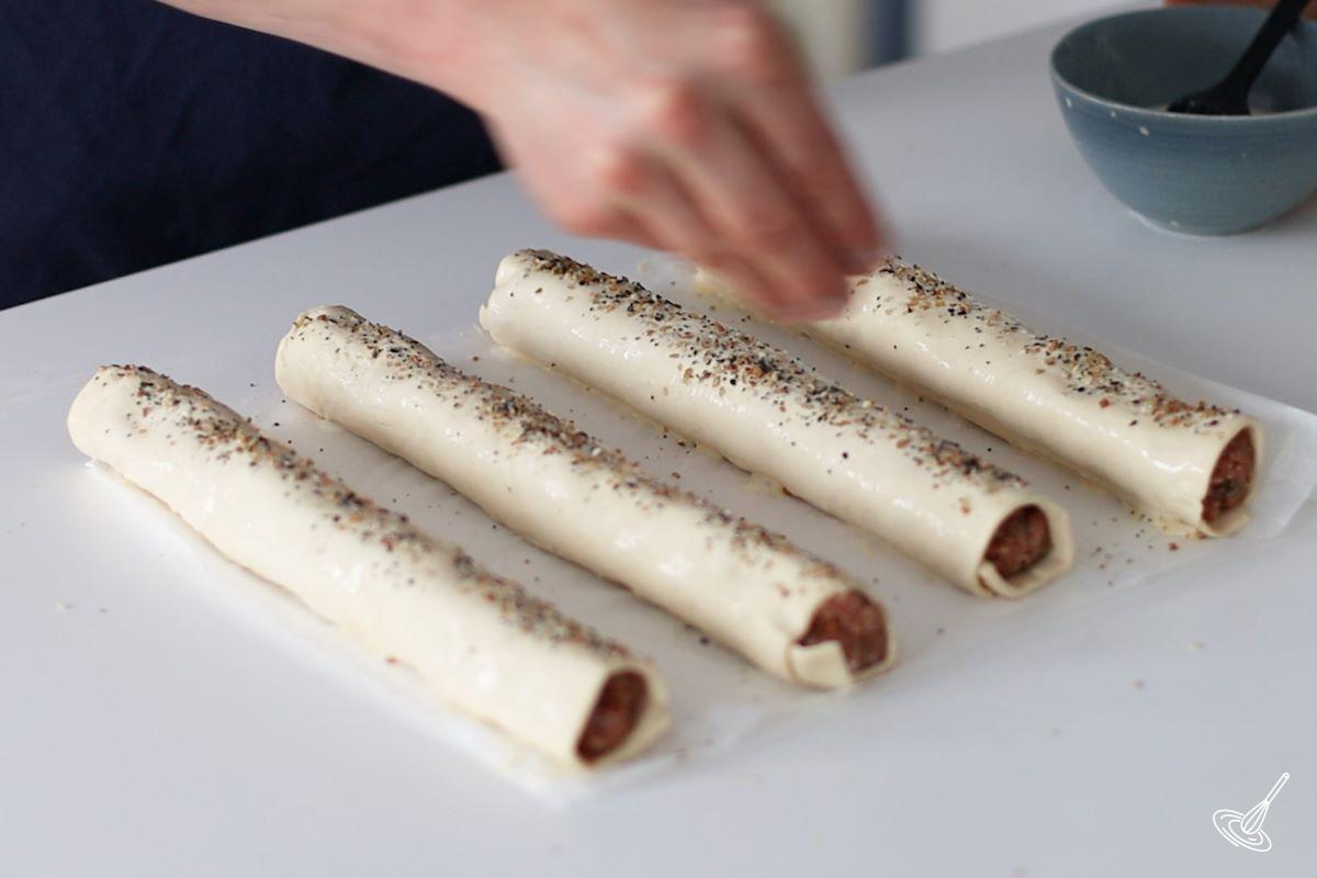 Someone sprinkling everything bagel seasoning on top of prepared french onion sausage rolls.