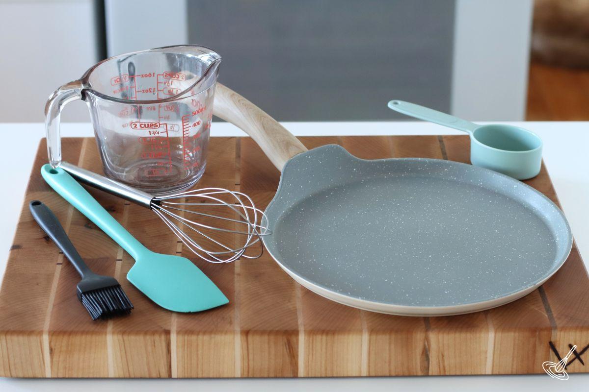 Tools to make crepes on a wood board, including a crepe pan, and mixing tools. 