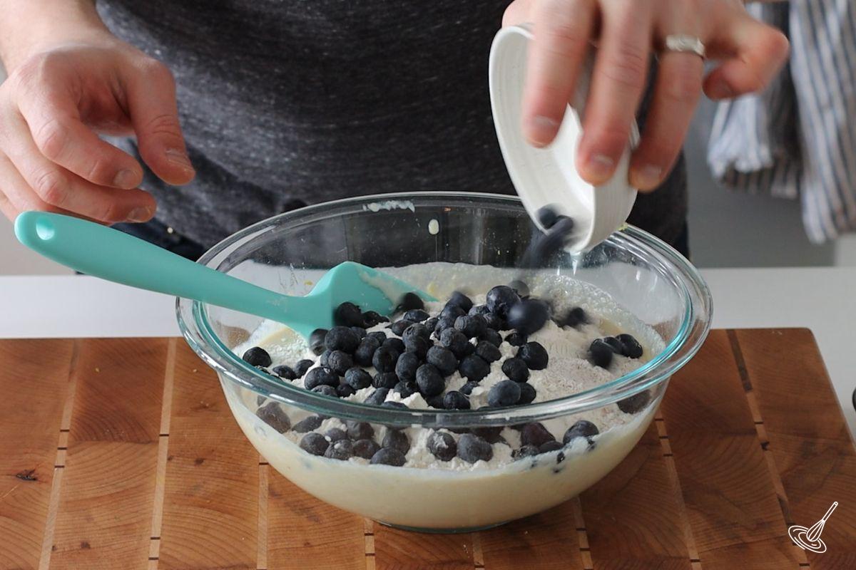 Someone adding blueberries to a bowl of muffin batter.
