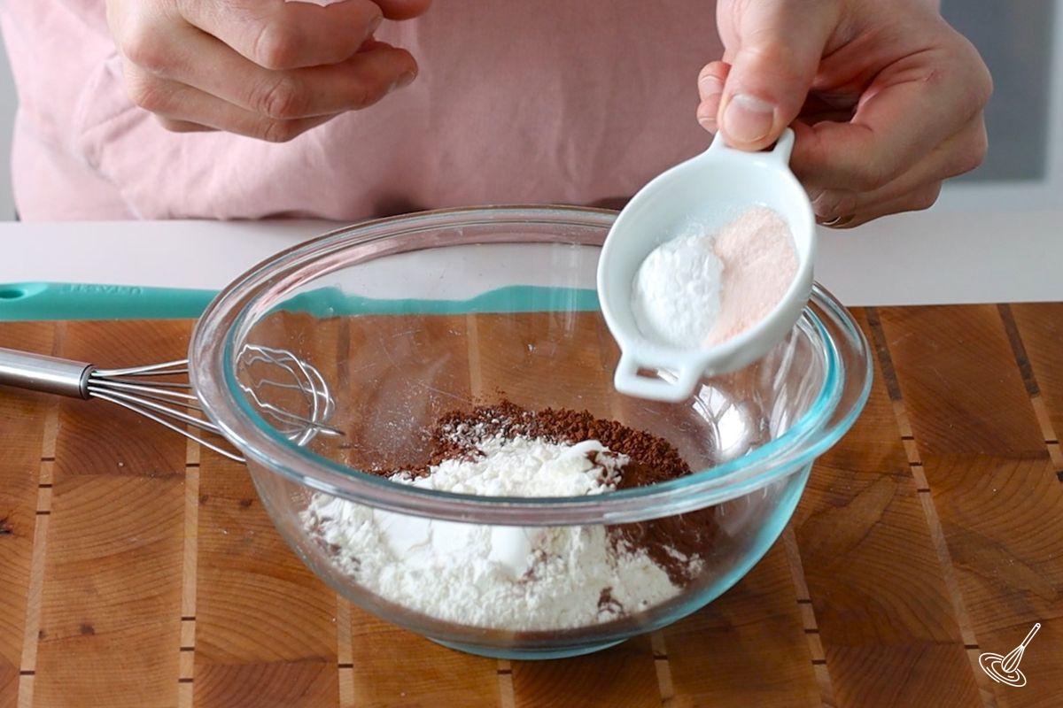 Someone adding salt to a large bowl of flour with cocoa powder.