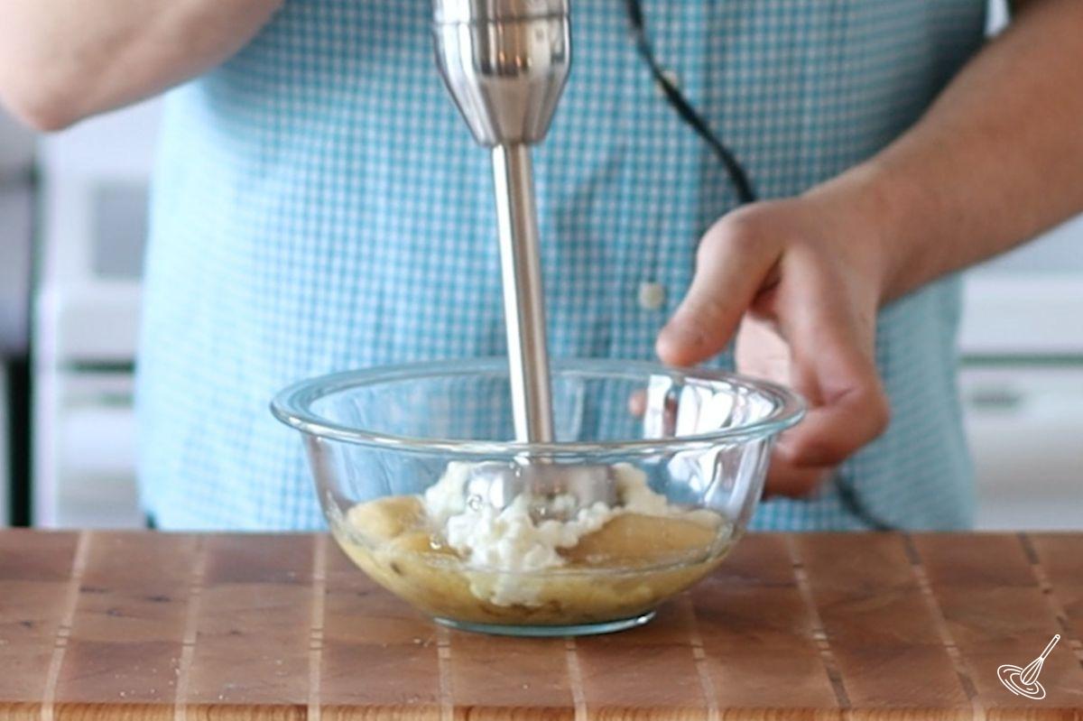 Someone using a stick blender to puree cottage cheese with bananas.