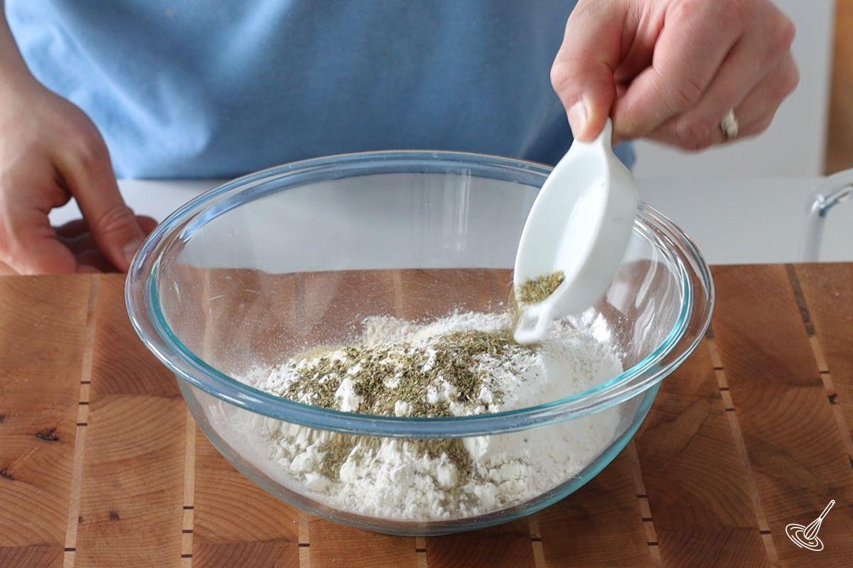 Someone adding dry herbs to a flour mixture in a bowl.