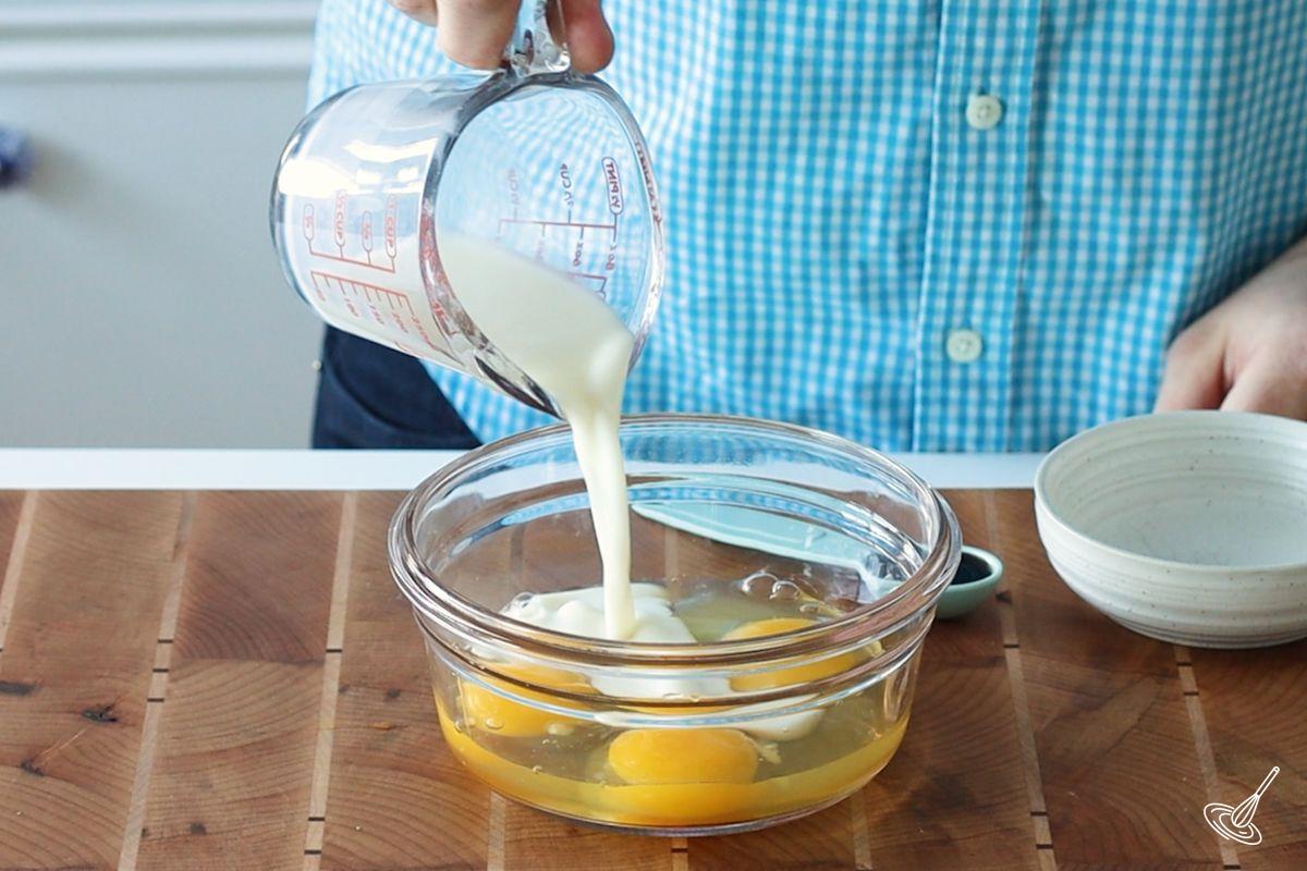 Someone pouring milk into a bowl with eggs. 