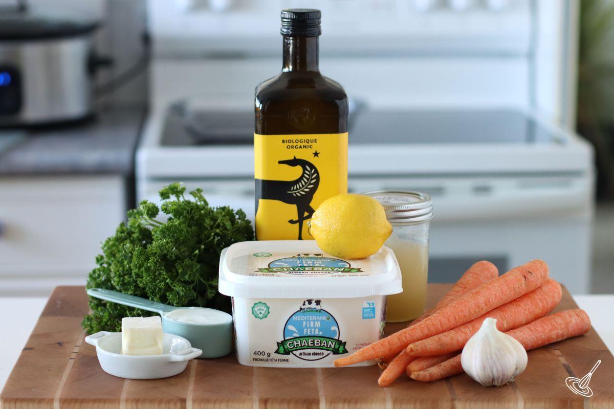 Ingredients on a wooden board, including carrots, feta, olive oil and fresh parsley.