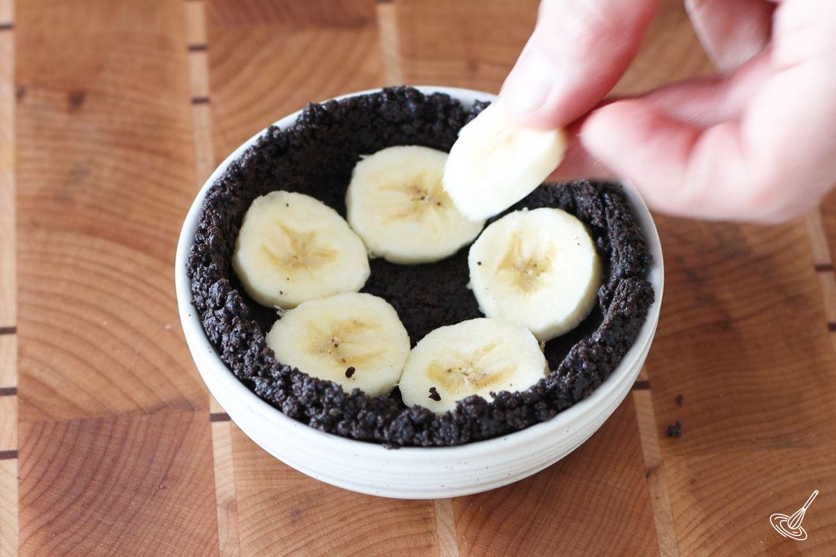 Someone placing slices of banana on the bottom of a small chocolate tart crust.