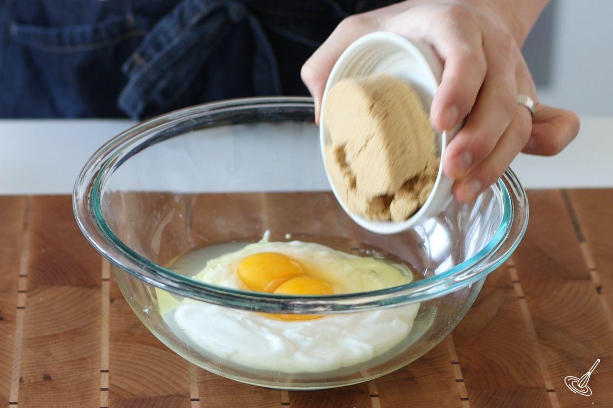 Someone adding brown sugar to a large bowl containing cottage cheese and eggs. 
