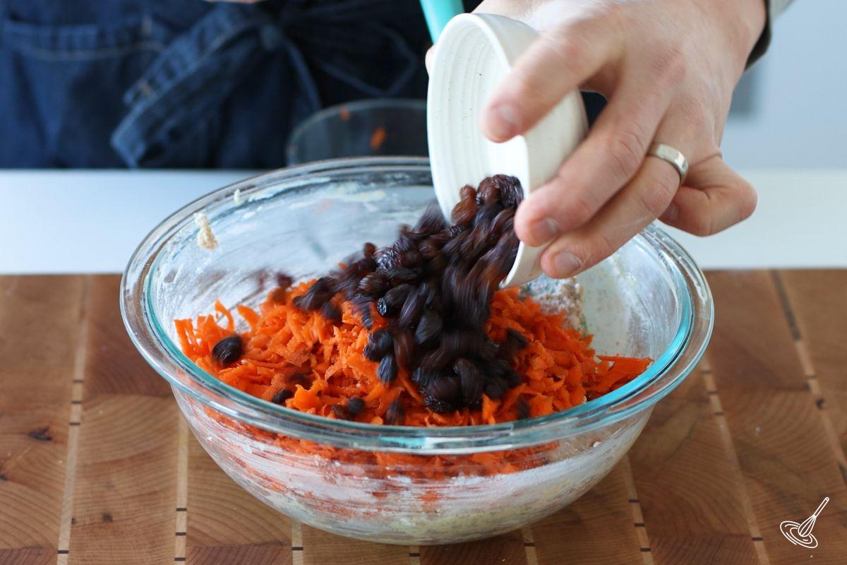 Someone adding raisins and grated carrots to a bowl of muffin batter.