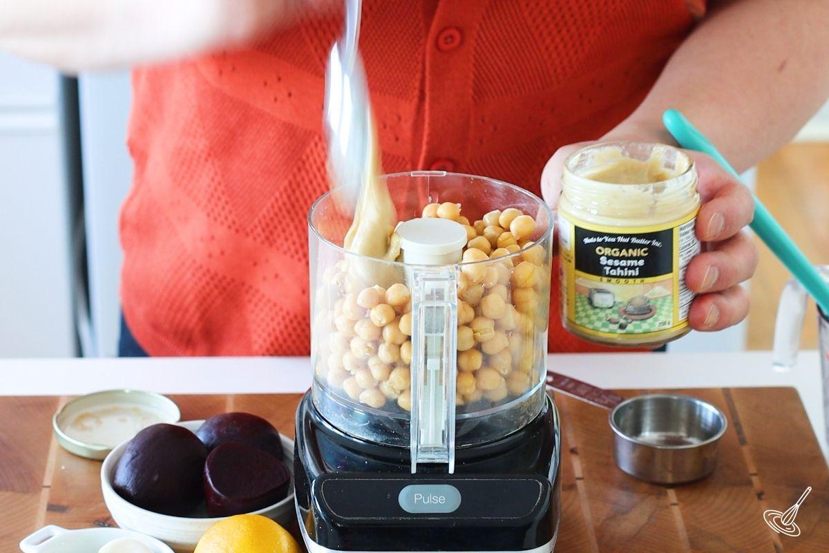 Someone adding tahini to a food processor containing chickenpeas. 