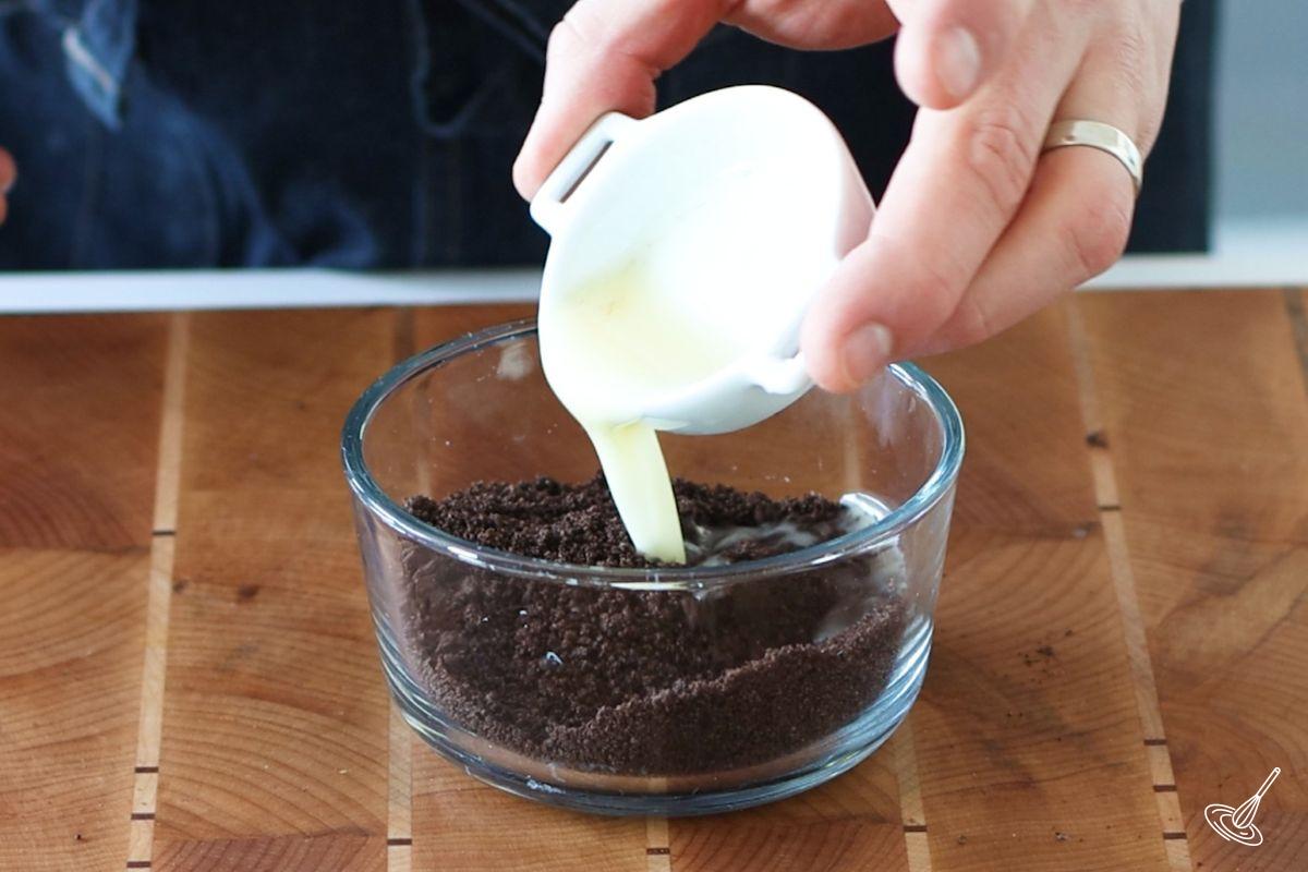 Someone pouring melted butter into a small container of chocolate cookie crumbs.