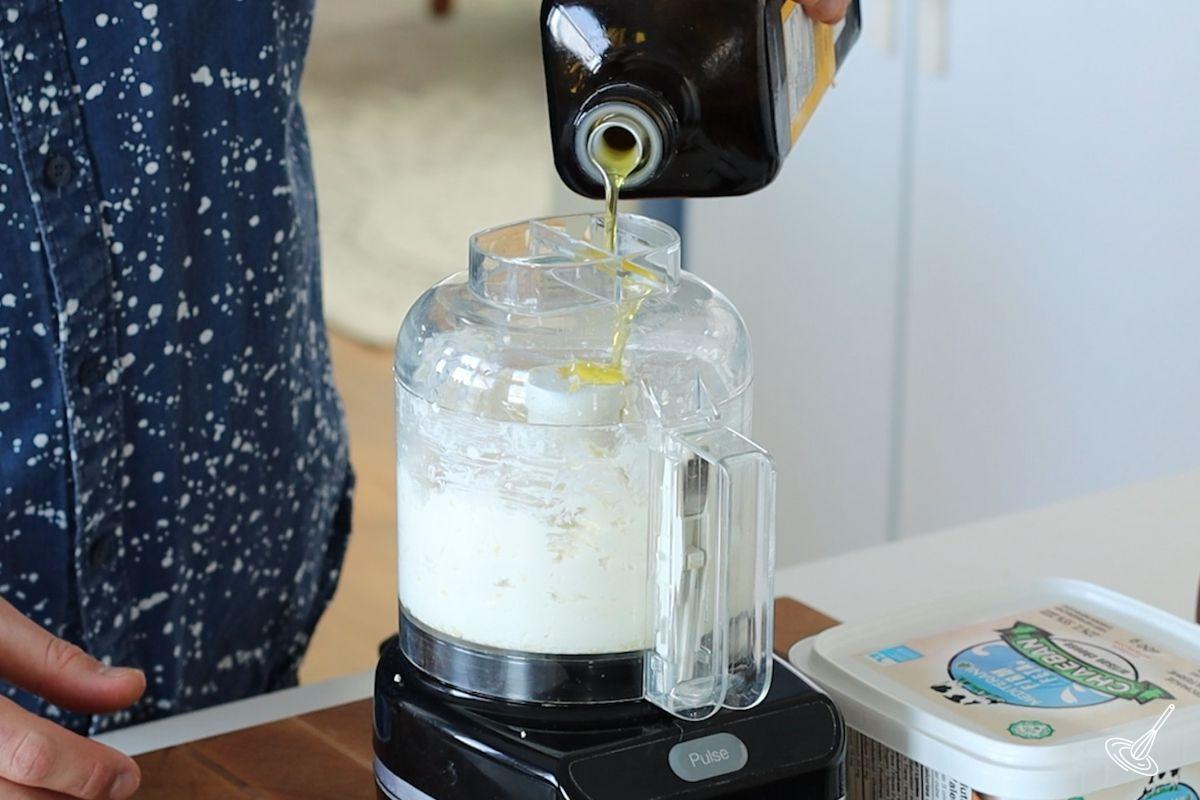 Someone pouring olive oil into a food processor filled with whipped feta.