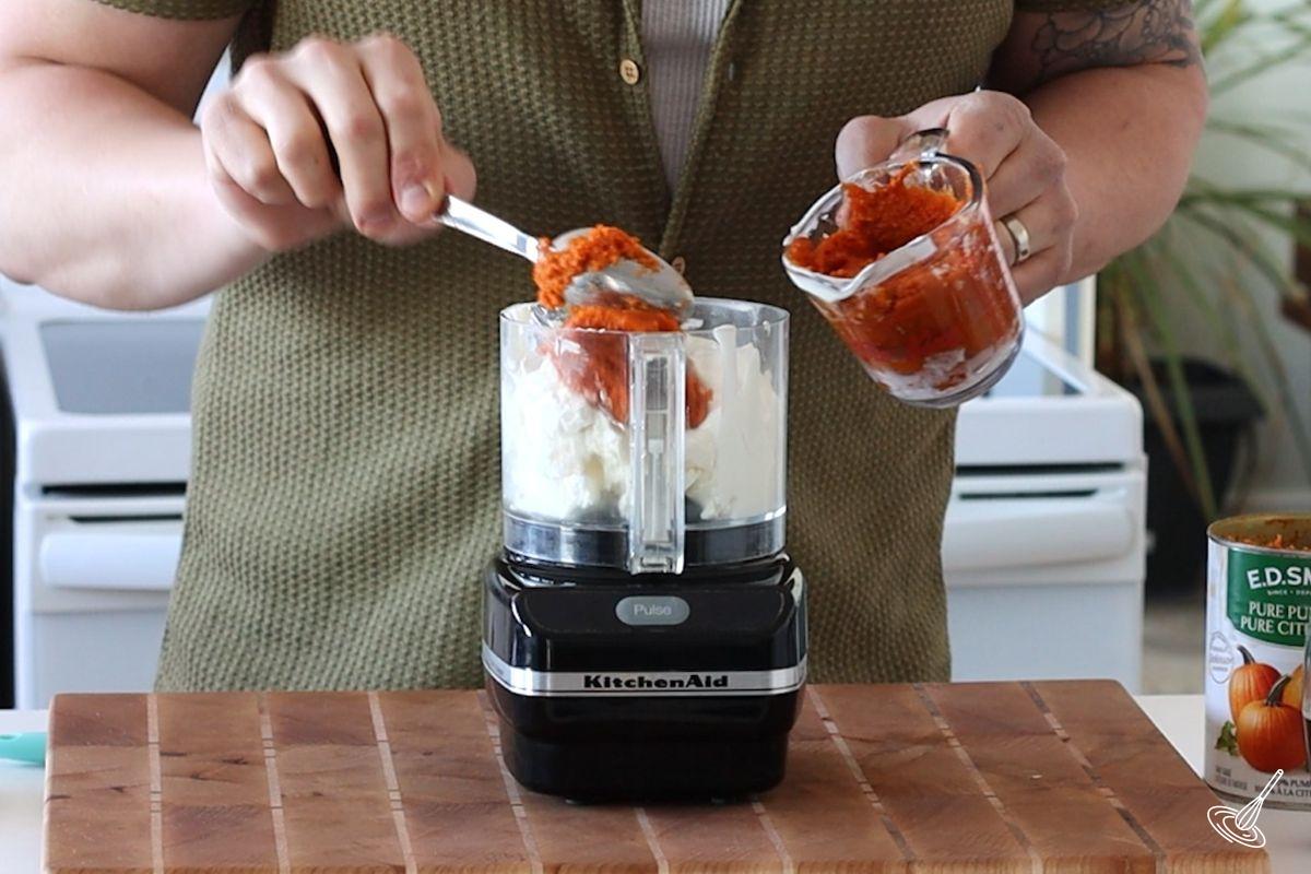 Someone adding pumpkin puree to a food processor.