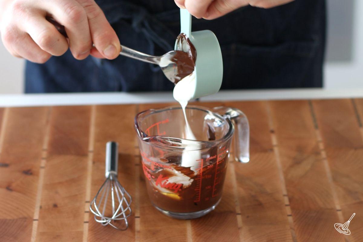 Someone adding sour cream to a small measuring cup of melted chocolate. 