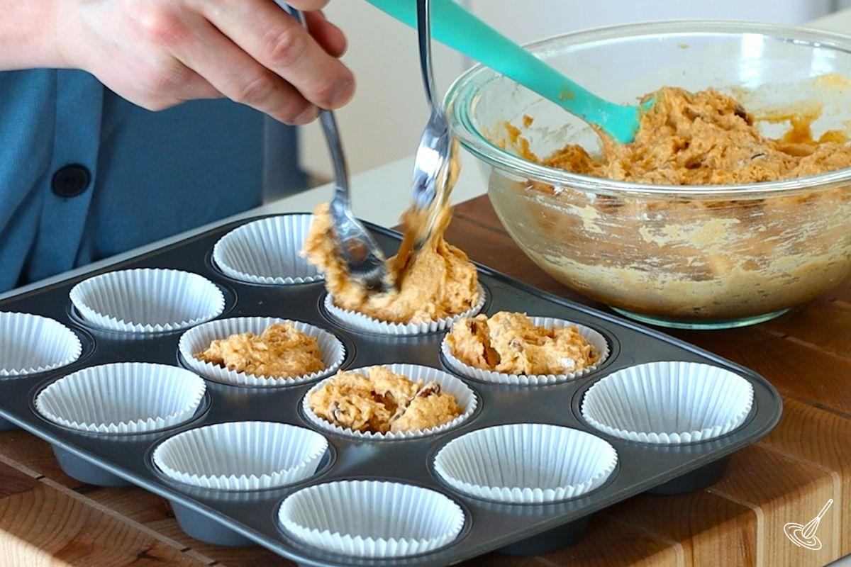 Someone using two large spoons to place pumpkin cottage cheese muffins batter into muffins tins.