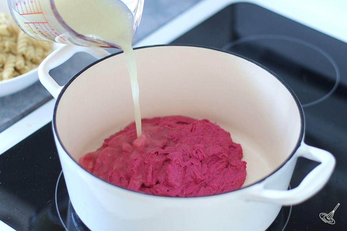 Someone pouring pasta water in a large sauce pan with beet and feta sauce. 