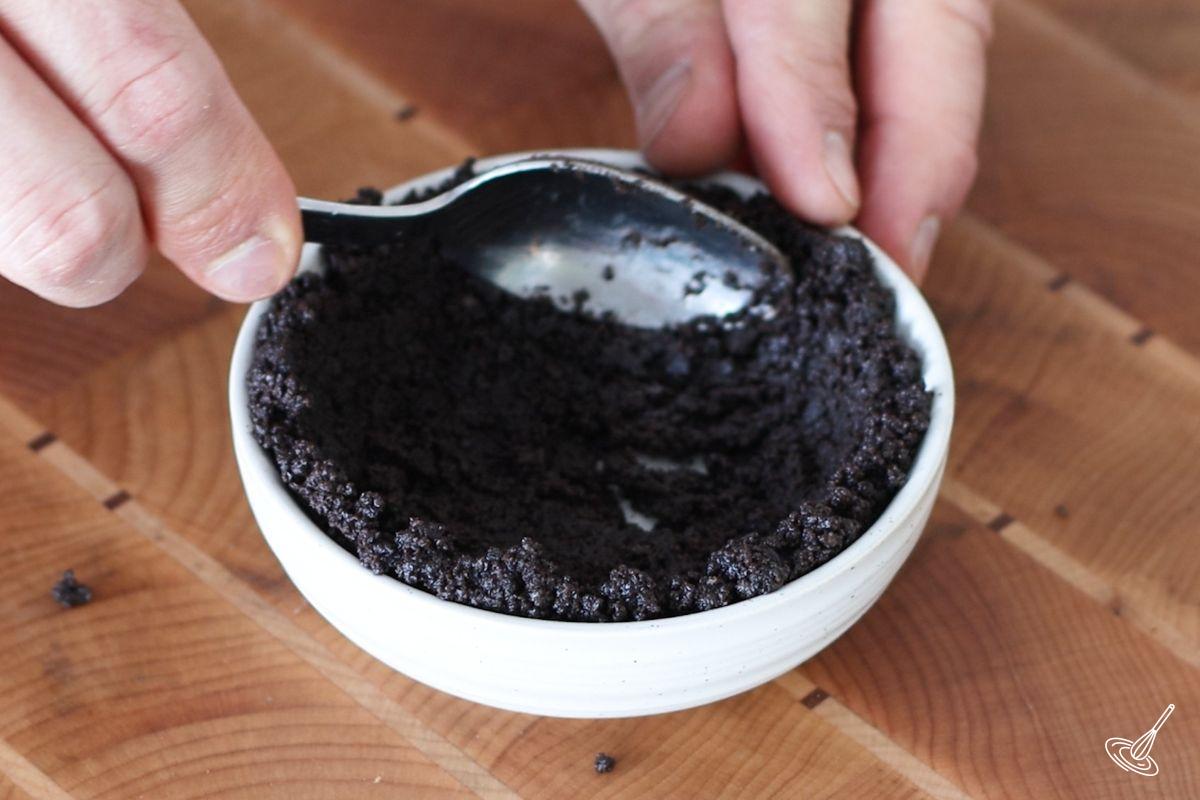 Someone using the back of a spoon to press a chocolate cookie crumb mixture on the sides of a small ramequin.