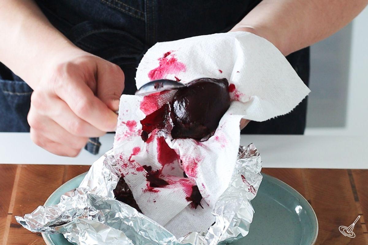 Someone holding a roasted beet with a paper towel and using a spoon to remove the skin. 