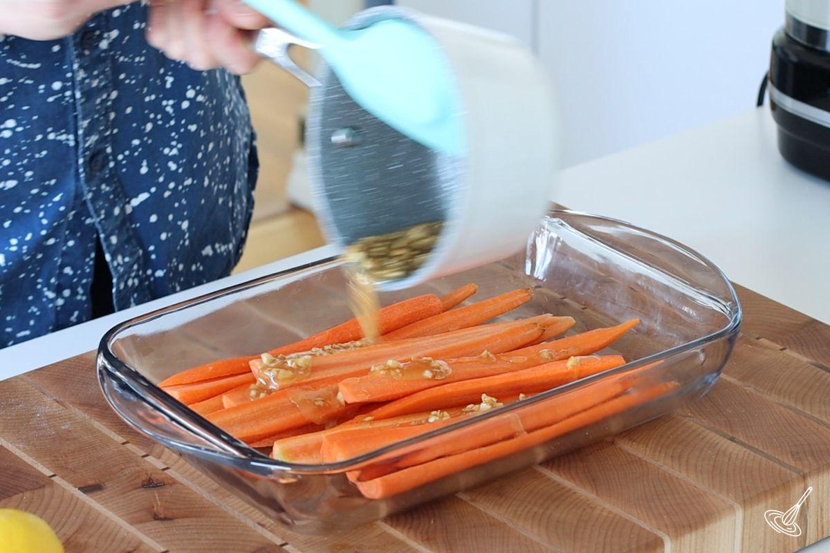 Someone pouring a honey glaze on top of prepared carrot in a baking dish.