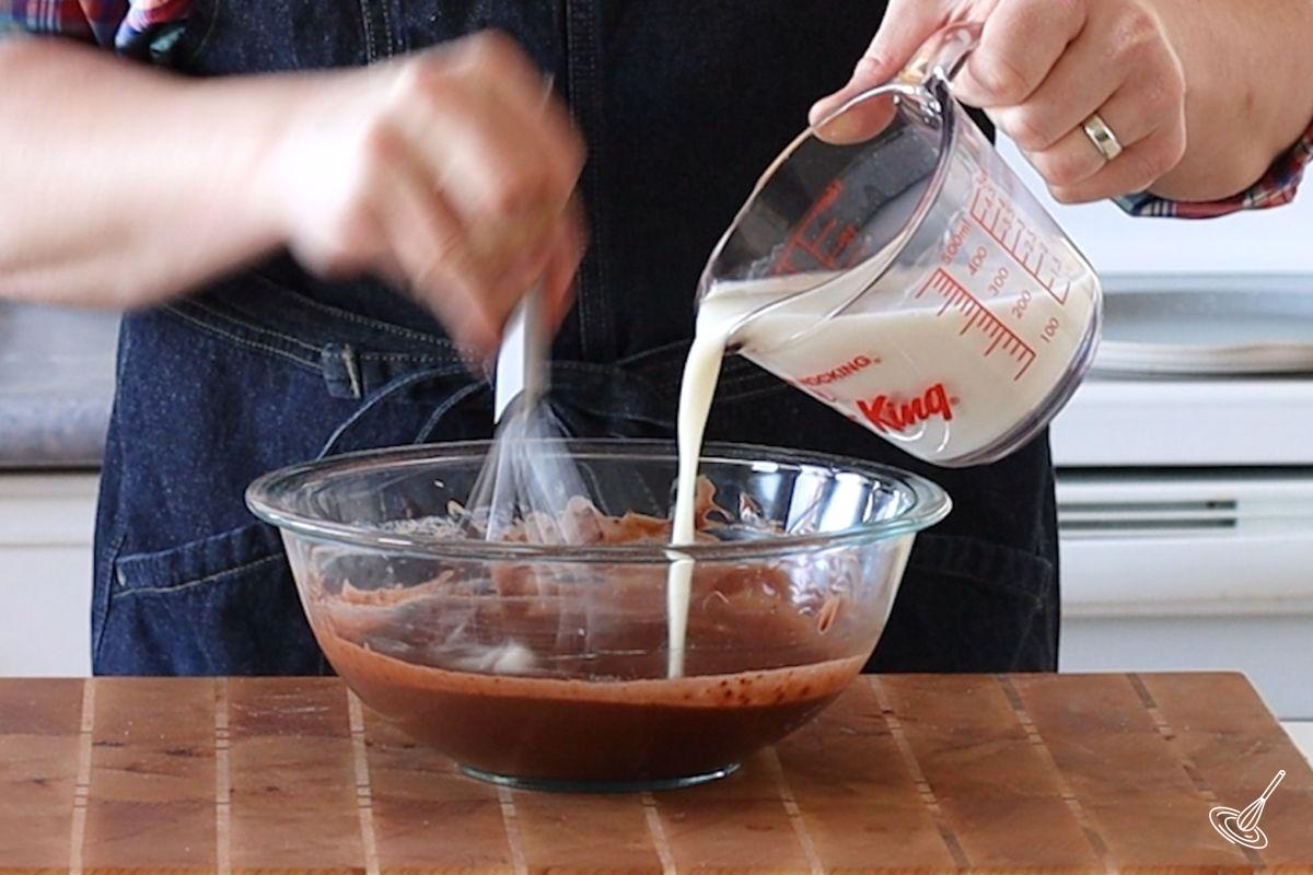 Someone slowly pouring in milk into a bowl of chocolate batter. 