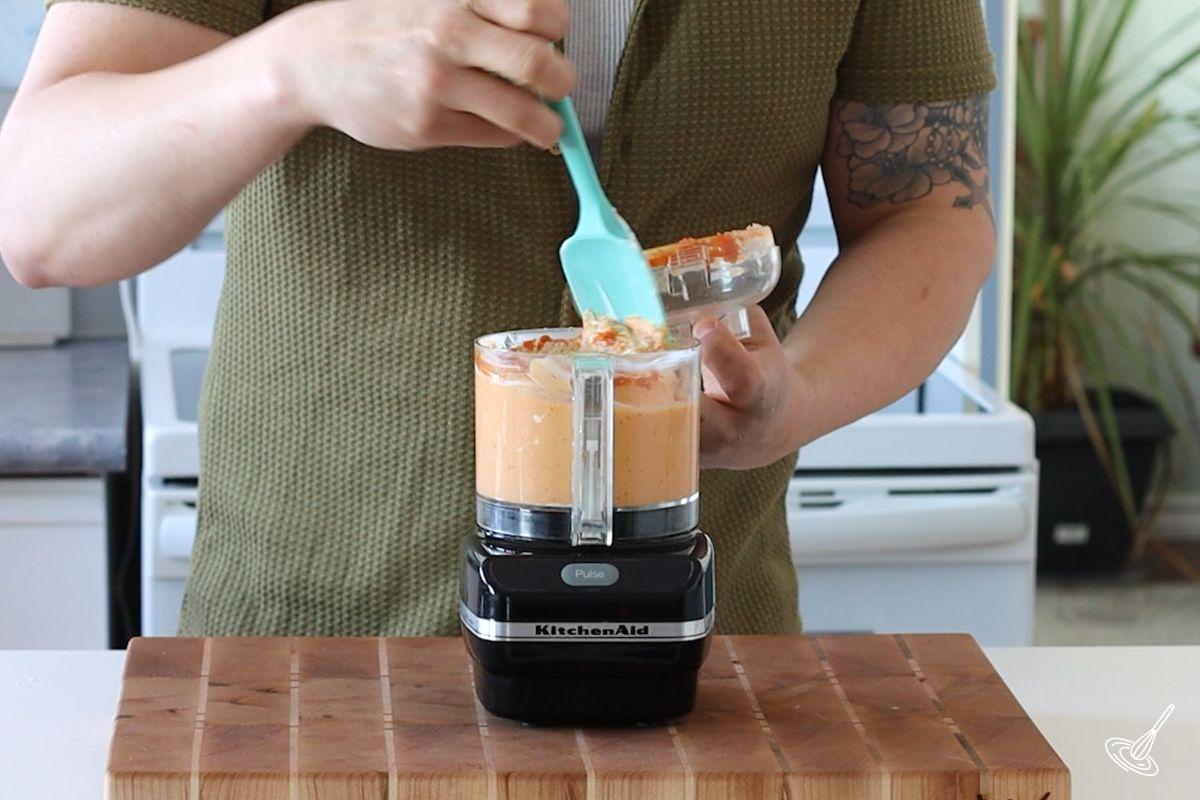 Someone using a rubber spatula to scrape down the sides of a food processor. 